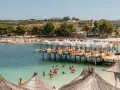 Słoneczny dzień na Ksamil Beach. Ludzie kąpią się w turkusowej wodzie, relaksują się na leżakach pod białymi parasolami i na drewnianych pomostach.