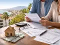Pareja revisa papeles y calcula los gastos de compra de vivienda segunda mano. Casa de madera, llaves y dinero sobre la mesa.