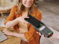 Uśmiechnięta kobieta zbliża telefon do terminala płatniczego, pokazując, jak płacić telefonem. Szybko i wygodnie.