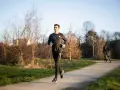 Un jeune homme s'entraîne à la course à pied sur un chemin ensoleillé, entouré d'arbres dénudés. Un cycliste passe en arrière-plan.