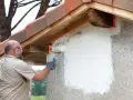 Un homme applique une peinture sur mur béton extérieur avec un rouleau. La rénovation progresse sous le toit de tuiles.