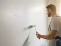Un peintre applique un crépi au rouleau sur un mur blanc. La lumière du soleil éclaire la scène.