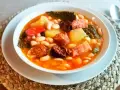 Un reconfortante cocido montañés, rebosante de alubias blancas, patatas, chorizo, morcilla y tocino, servido en un cuenco blanco.