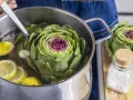 Artischocke zubereiten: Eine ganze Artischocke wird in einem Topf mit Wasser und Zitronenscheiben gekocht. Daneben liegen weitere Artischocken und ein Glas Salz.