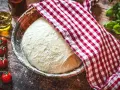Frischer Pizzateig ruht in einer Schüssel, bedeckt mit einem rot-weiß karierten Tuch. Zutaten für ein Pizzateig Rezept wie Tomaten und Öl liegen bereit.