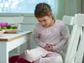 Mała dziewczynka siedzi przy stole, trzymając się za brzuch. Widać, że cierpi z powodu bólu brzucha u dziecka.