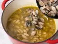 Alubias cremosas con almejas en una olla roja, añadiendo más almejas de una sartén.