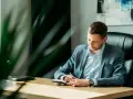 Mężczyzna w garniturze siedzi przy biurku i pisze w notatniku.
