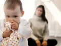 Kleines Baby hält sich an einem Tuch fest, wirkt schüchtern. Im Hintergrund sitzt eine unscharfe Frau.