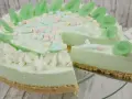 Eine helle, cremige Philadelphia Torte Waldmeister mit bunten Marshmallows und grünen Blättern verziert. Ein Stück wird gerade herausgeschnitten.