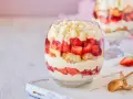 Postre en vaso con capas de crema, fresas y bizcocho, cubierto con trozos de chocolate blanco.