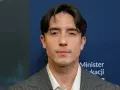 Dawid Kwiatkowski w szarym garniturze na tle niebieskiej ściany z napisami.