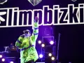 Fred Durst z Limp Bizkit na koncercie, w neonowej kurtce, z ręką w górze. Tło to logo zespołu.