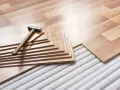 Tablas de suelo laminado apiladas con martillo y lápiz, listas para aislar suelo frío sobre una base acolchada.