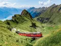 A red Brienz Switzerland train winds through lush green mountains, with a stunning blue lake in the distance.