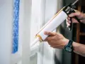 Ręce trzymają pistolet do silikonu, aplikując go na ramę okna. To pokazuje, co to jest silikon – materiał uszczelniający.