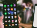 Dłoń trzyma smartfon z widocznym ekranem pełnym ikon aplikacji, w tym ikoną wiadomości SMS.