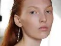 Primo piano di una giovane donna con capelli rossi e occhi azzurri, un neo della bellezza sul viso. Indossa un orecchino pendente.