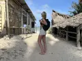 Kobieta z plecakiem na piaszczystej plaży, zastanawia się co zabrać na Zanzibar. W tle chatki z palmowymi dachami i budynek z kamienia.