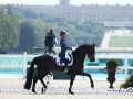 Un cavalier dressage français, en tenue bleue et blanche, monte un cheval noir dans une carrière. Le château de Versailles est visible en arrière-plan.