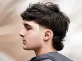 Młody mężczyzna z kręconymi włosami i modną fryzurą mullet. Krótsze boki i dłuższe włosy z tyłu nadają mu charakteru.