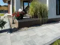 Nowy, tani chodnik przed domem z szarych płyt, ozdobiony donicami z kwiatami i trawami, tworzy estetyczne wejście.
