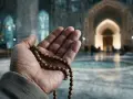 Eine Hand hält eine Gebetskette, ein Symbol des christlichen Glaubensbekenntnisses, vor einer Moschee.