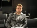 Marcin Hycnar w szarej marynarce i bluzie z kapturem siedzi na czarnym krześle, wpatrując się wprost przed siebie.