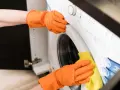 Hände in orangen Handschuhen reinigen die Waschmaschine. Ein gelber Lappen wischt über die Oberfläche, um sie sauber zu halten.