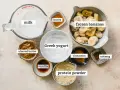 Składniki do zrobienia odżywki białkowej: mleko, jogurt grecki, mrożone banany, masło orzechowe, miód, wanilia, cynamon, gałka muszkatołowa i proszek białkowy.