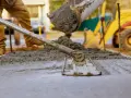 Pracownik rozprowadza świeży beton c12/15 za pomocą łaty. W tle widać koparkę i plac budowy.