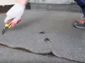 Ręka w rękawicy tnie nożem papę na dachu, przygotowując do naprawy. Widoczne uszkodzenia i pęknięcia.