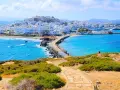 A stone path leads to a causeway connecting to the whitewashed town of Naxos, Greece, with vibrant blue waters lapping at the shore.