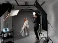 Profesjonalna fotografia studyjna: kobieta w jasnym garniturze pozuje na białym sześcianie, fotograf robi jej zdjęcie.