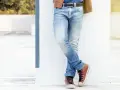 Mężczyzna w niebieskich jeansach męskich i brązowej kurtce stoi oparty o białą ścianę.