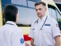 Ratownik medyczny w białej koszuli ze stetoskopem i naszywkami omawia sytuację z koleżanką przy karetce.