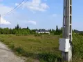 Betonowe słupy energetyczne stoją na tle zieleni i nieba. W oddali widać domki jednorodzinne.