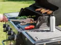 Ręka z szczypcami obraca na grillu kawałki sera halloumi, obok pieczone pomidory i papryczki.