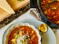 Aksamitna zupa minestrone z kawałkami warzyw i startym parmezanem, podana w ozdobnej misce. Obok garnek z zupą i kawałek sera na desce.