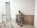 Budowlaniec montuje płyty regipsowe na stelażu. Obok stoi drabina.