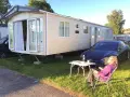 En person njuter av solen vid ett campingbord utanför en husvagn. En Tesla parkerad bredvid. Camping Timrå.