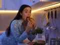 Kobieta w piżamie z gwiazdkami je kanapkę, choć czuje, że jest głodna, ale nie ma apetytu. Na blacie leży też pizza i burger.