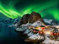 Grön norrsken dansar över snöklädda berg och röda fiskestugor vid en vik i Norge.