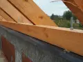 Drewniana belka, czyli murłata, jest przykręcona do betonowego wieńca za pomocą śrub. W tle widać fragmenty dachu i drzewa.