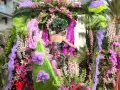 Una figura con un vestido floral y un tocado verde, rodeada de exuberantes flores rosas y moradas, participa en el **carnaval de Nice**.