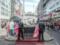 Zwei Männer in Uniformen mit US-Flaggen stehen am Checkpoint Charlie in Berlin. Touristen und ein McDonald's im Hintergrund.