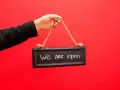 Mano con chaqueta negra sostiene un cartel que dice "We are open", indicando la apertura de un negocio, similar a los tipos de licencia de actividad.