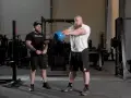 Man med skägg utför kettlebell swing varje dag i gym. Bredvid står en tränare.