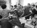 Un grupo de jóvenes y soldados interactúan tras una barricada de piedras en Argelia. Un hombre con uniforme militar sirve bebida.