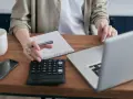 Persona revisa cuentas en su portátil, calculadora y libreta. Podría estar gestionando la hacienda pública deudora por IVA.
